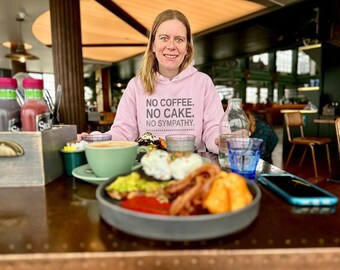 May include: A light pink hooded sweatshirt featuring the text NO COFFEE. NO CAKE. NO SYMPATHY. printed in dark letters on the front. The garment is worn by a person sitting at a table with a plate of food, a green coffee cup, and a glass of water.