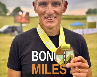 May include: A black t-shirt with the white and orange text "BONUS MILES" and a dashed line underneath. A yellow lanyard holds a circular medal featuring a mountain graphic in green, white, and tan colors. The medal is held up by a hand.