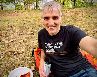 May include: A black short-sleeved t-shirt featuring the white text "THAT'S THE HARD PART DONE." followed by the orange text "MORE HARD PARTS TO COME." with a dotted line underneath, worn by a person with short gray hair.