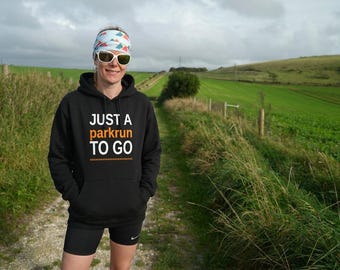 May include: A black hooded sweatshirt featuring the white and orange text "JUST A parkrun TO GO" with a dotted line underneath. The hoodie includes a front kangaroo pocket and matching black drawstrings, paired with black athletic shorts.