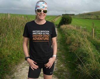 May include: A black short-sleeved t-shirt featuring the white and orange text "ANOTHER MARATHON. ANOTHER ULTRA. ANOTHER CRAZY ADVENTURE." with a dashed line underneath, paired with black athletic shorts, a patterned headband, and sunglasses.