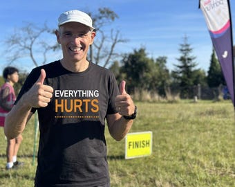 May include: A black short-sleeved t-shirt featuring the white text "EVERYTHING" above the orange text "HURTS" with a dashed line underneath. The shirt is worn with a white baseball cap, and the design is printed clearly on the front chest area.
