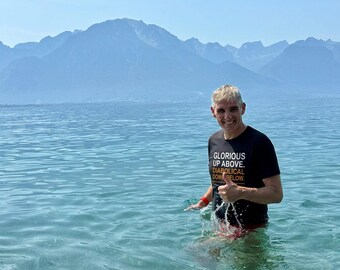 May include: A black short-sleeved t-shirt featuring the white and orange text GLORIOUS UP ABOVE. DIABOLICAL DOWN BELOW. The shirt is worn by a person standing in clear blue water with a large mountain range visible in the background.