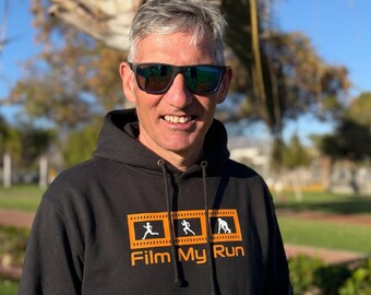 May include: A black hooded sweatshirt featuring a graphic of three white runner silhouettes inside orange film strip frames, with the text "Film My Run" printed in orange below the graphic. The hoodie has a drawstring hood and long sleeves.