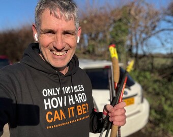 May include: A black hooded sweatshirt featuring white and orange text that reads ONLY 100 MILES HOW HARD CAN IT BE? The garment includes a drawstring hood and long sleeves, worn while holding a pair of trekking poles with cork handles and yellow accents.