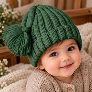 May include: A dark green knit beanie featuring a ribbed texture and a large, matching yarn pom-pom attached to the side. The hat has a folded cuff design and is crafted from a thick, soft-looking yarn material with a consistent deep green color.