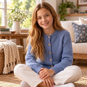 May include: A periwinkle blue long-sleeve knit cardigan featuring a crew neckline, ribbed cuffs, and a front button closure with five light-colored buttons. The garment is worn over white pants, showcasing a textured knit pattern throughout.