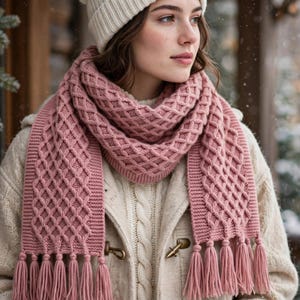 May include: A dusty rose knitted scarf featuring a textured diamond cable pattern and long fringe tassels at both ends. The scarf is worn over a cream-colored cable knit sweater and is paired with a matching cream-colored knit beanie hat.