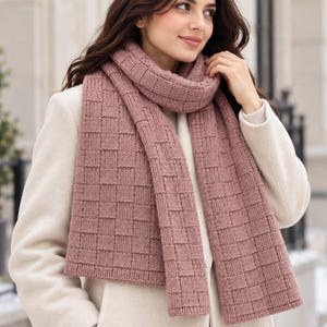 May include: A dusty rose knitted scarf featuring a textured basketweave pattern. The scarf is draped around the neck and hangs down the front, layered over a cream-colored coat. The knit construction displays a repeating square grid design.