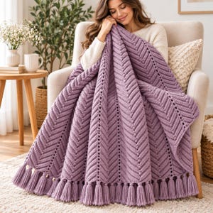 Könnte beinhalten: Eine lavendelfarbene Strickdecke mit einem markanten Fischgräten- oder Chevron-Muster. Die Decke ist am unteren Rand mit einer Reihe passender lavendelfarbener Garnquasten versehen, die ein dekoratives Fransendetail bilden.
