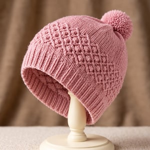 May include: A dusty pink knit beanie featuring a ribbed brim and a textured diamond pattern with small raised bobbles across the main body. The hat is topped with a matching round pom-pom and displays a consistent knit construction throughout.