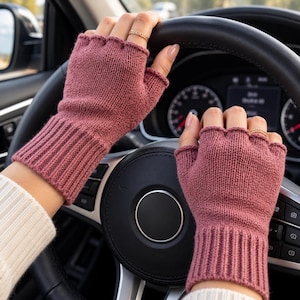 May include: A pair of dusty rose fingerless gloves featuring a ribbed knit cuff and a scalloped edge at the knuckles. The gloves are made of a soft knit material and are worn while gripping a black leather steering wheel inside a vehicle.