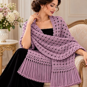 May include: A lavender crochet shawl featuring an intricate diamond-patterned openwork design, a scalloped border, and long, straight fringe detailing along the bottom edges. The wrap is draped over a black garment, showcasing the textured yarn work.