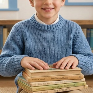 May include: A blue long-sleeved knit sweater with a crew neckline worn over a white collared shirt. The sweater features a ribbed collar, cuffs, and hem, and is worn by a person with hands resting on a stack of five hardcover books.