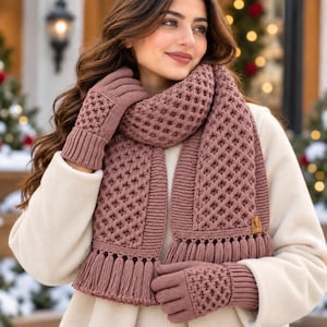 May include: A dusty rose knitted scarf and matching gloves set featuring a textured waffle stitch pattern. The scarf includes a ribbed border and a row of fringe tassels at the ends. A small square leather-like tag with the letter M is attached.