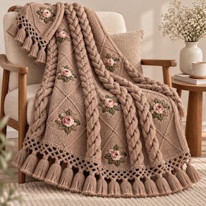Peut inclure: Un jeté de lit en tricot rose poudré avec un motif en losanges, des bandes verticales en torsades épaisses et des motifs de roses brodées avec des feuilles vertes. Les bords sont ornés d'une bordure en treillis et de longs pompons assortis.