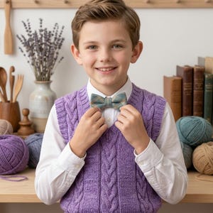 Può includere: Un gilet in maglia a trecce color lavanda con scollo a V, indossato sopra una camicia bianca a maniche lunghe con bottoni. Un papillon a quadri blu e beige è fissato al colletto, completando l'insieme a strati con un motivo lavorato a maglia.