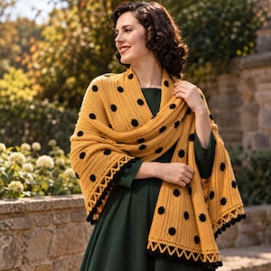 May include: A mustard yellow knitted shawl featuring a ribbed texture and small black floral-shaped appliques scattered across the fabric. The shawl is finished with a decorative triangular zigzag border and small black pom-poms along the bottom edge.
