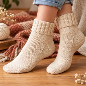 Könnte beinhalten: Ein Paar cremefarbene Strickstrümpfe mit geripptem Bündchen. Die Socken sind mit einer sichtbaren Fersennaht und einer gleichmäßigen Strickstruktur im Fuß- und Knöchelbereich gefertigt und reichen bis zur Mitte der Wade mit einem Umschlag.