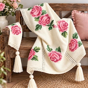 Op de afbeelding: Een crèmekleurige gebreide plaid met een kruissteekpatroon van roze rozen met groene bladeren. De deken heeft een decoratieve gehaakte rand en grote crèmekleurige kwastjes bevestigd aan de hoeken en randen van de stof.