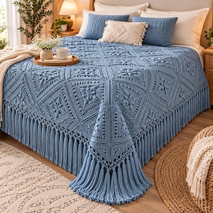 Puede incluir: Una colcha de ganchillo azul polvoriento con un patrón cuadrado repetitivo con motivos florales centrales y flecos largos en los bordes. La colcha está colocada sobre una cama con dos almohadas azules y una almohada central de color crema.