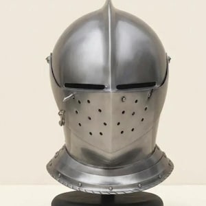 May include: A polished steel knight helmet featuring a central ridge, a horizontal eye slit, and a visor section with multiple ventilation holes. The helmet is secured to a circular black base and includes a metal neck guard with visible rivets.