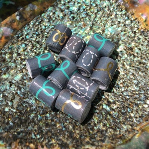 May include: A collection of small, rolled paper beads in various designs, including gold, teal, and white accents. The beads are arranged on a shimmering, iridescent surface, creating a visually appealing display.