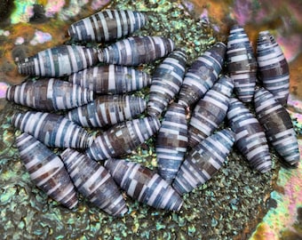 May include: A close-up of a group of handmade paper beads. The beads are a mix of white and gray with a striped pattern. The beads are arranged in a pile on a iridescent surface.