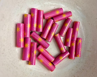 May include: A collection of pink wooden beads, each adorned with a yellow sun design. The beads are cylindrical and scattered in a neutral-toned bowl. The sun design is centered on each bead.