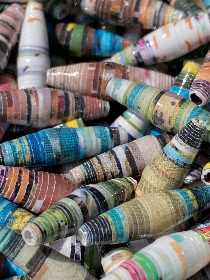 May include: A close-up of a pile of colorful, striped paper beads. The beads are made from rolled up pieces of paper and are all different lengths and widths. The beads are a mix of bright colors, including blue, green, red, yellow, and orange.