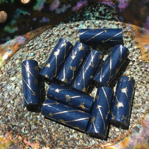 May include: A group of eight dark blue cylindrical beads with gold glitter and a gold arrow pattern. The beads are arranged in a pile on a textured iridescent surface.