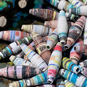 May include: A close-up of a collection of colorful, striped, paper beads. The beads are made from rolled up strips of paper and are arranged in a pile. The beads are a variety of colors, including blue, green, red, yellow, and white.