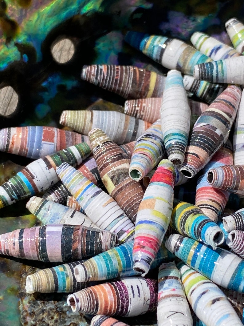 May include: A close-up of a collection of colorful, striped, paper beads. The beads are made from rolled up strips of paper and are arranged in a pile. The beads are a variety of colors, including blue, green, red, yellow, and white.