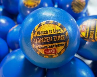 May include: A blue foam stress ball featuring a circular yellow and black hazard-style sticker that reads Watch It Live! DANGER ZONE Starring John Ordover with a YouTube logo. The ball has a smooth, rounded surface and is part of a group of identical items.