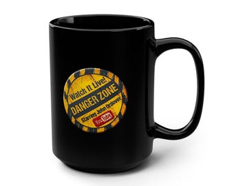 May include: A black ceramic mug featuring a circular yellow and black hazard-striped graphic. The text reads Watch It Live! DANGER ZONE Starring John Ordover with a red YouTube logo. The design has a distressed, weathered appearance.