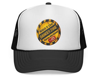 May include: A white and black trucker cap featuring a circular graphic on the front panel. The graphic has a yellow and black hazard stripe background with the text Watch It Live! DANGER ZONE Starring John Ordover and a YouTube logo.