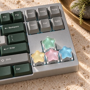 May include: A silver mechanical keyboard featuring dark green and grey rectangular keycaps, with four custom star-shaped keycaps in translucent yellow, green, pink, and blue colours. The keyboard includes standard keys labelled Backspace, Enter, Shift, Ctrl, Insert, Home, PgUp, Delete, End, and PgDn.
