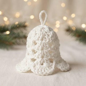May include: A white crocheted bell ornament featuring an intricate openwork stitch pattern and a small loop at the top for hanging. The bell has a flared, scalloped bottom edge and is crafted from soft white yarn, creating a textured appearance.