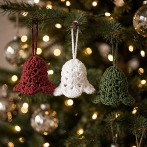 May include: Three crochet bell ornaments hanging from a tree, featuring one deep red, one white, and one dark green bell. Each bell is crafted with a textured, open-stitch pattern and includes a matching yarn loop for hanging on a branch.