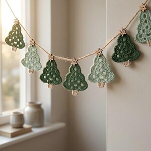 May include: A garland featuring crocheted Christmas trees in alternating shades of sage and forest green, each with a small beige trunk. The trees are strung on twine and accented with small, laser-cut wooden snowflake ornaments.