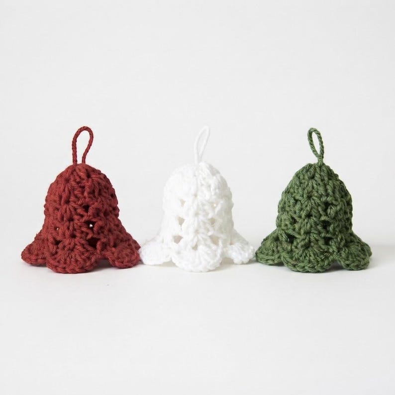 May include: Three crocheted bell ornaments in dark red, white, and olive green. Each bell features a textured, open-stitch pattern with a scalloped bottom edge and a small yarn loop at the top for hanging, arranged in a horizontal row.