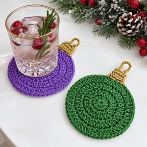 May include: Two circular crochet coasters shaped like Christmas ornaments, one purple and one green, each featuring a gold-colored crocheted cap with a loop at the top. A clear glass filled with ice, cranberries, and a rosemary sprig sits on the purple coaster.