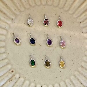 Op de afbeelding: Een verzameling van tien kleine zilverkleurige hangers, elk voorzien van een enkele ovaalvormige edelsteen in diverse kleuren waaronder transparant, rood, paars, blauw, roze, groen, bruin en geel, opgehangen aan een kleine zilveren ring.
