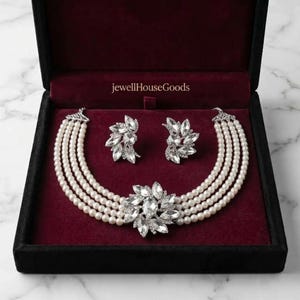 May include: A four-strand pearl necklace and matching pair of earrings featuring clear marquise-cut crystals in a floral arrangement. The jewelry set is displayed in a black box with a deep red velvet interior, marked with the text jewellHouseGoods.