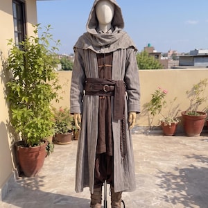May include: A full-length hooded cloak in a textured grey fabric worn over a dark brown tunic and trousers. A wide brown leather belt with a metal buckle is cinched at the waist. The outfit is completed with light brown boots on a mannequin.