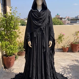 May include: A floor-length black hooded cloak featuring a layered, distressed fabric design with raw, frayed edges. The garment includes a deep hood, long sleeves, and a voluminous, flowing skirt that pools at the base, creating a textured appearance.