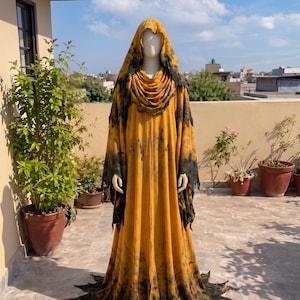 May include: A floor-length hooded robe featuring a mustard yellow and black tie-dye pattern. The garment includes a draped cowl neckline, long sleeves with jagged, distressed-style edges, and a hemline finished with pointed, flame-like fabric cutouts.