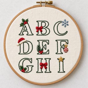 May include: A circular wooden embroidery hoop containing white fabric with hand-stitched letters A through I in dark green thread. Each letter is decorated with festive elements including holly, a red bow, a snowflake, a Santa hat, and ornaments.