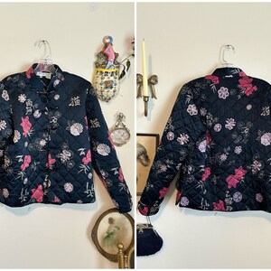 May include: A navy blue quilted jacket with a floral pattern in pink and white. The jacket has a mandarin collar and frog closures. The design includes Chinese characters. The jacket is displayed on a hanger.
