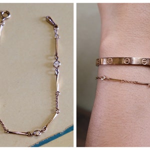 May include: Two gold bracelets on a wrist. The top bracelet is a thick band with a repeating pattern of circles. The bottom bracelet is a delicate chain with small round stones.